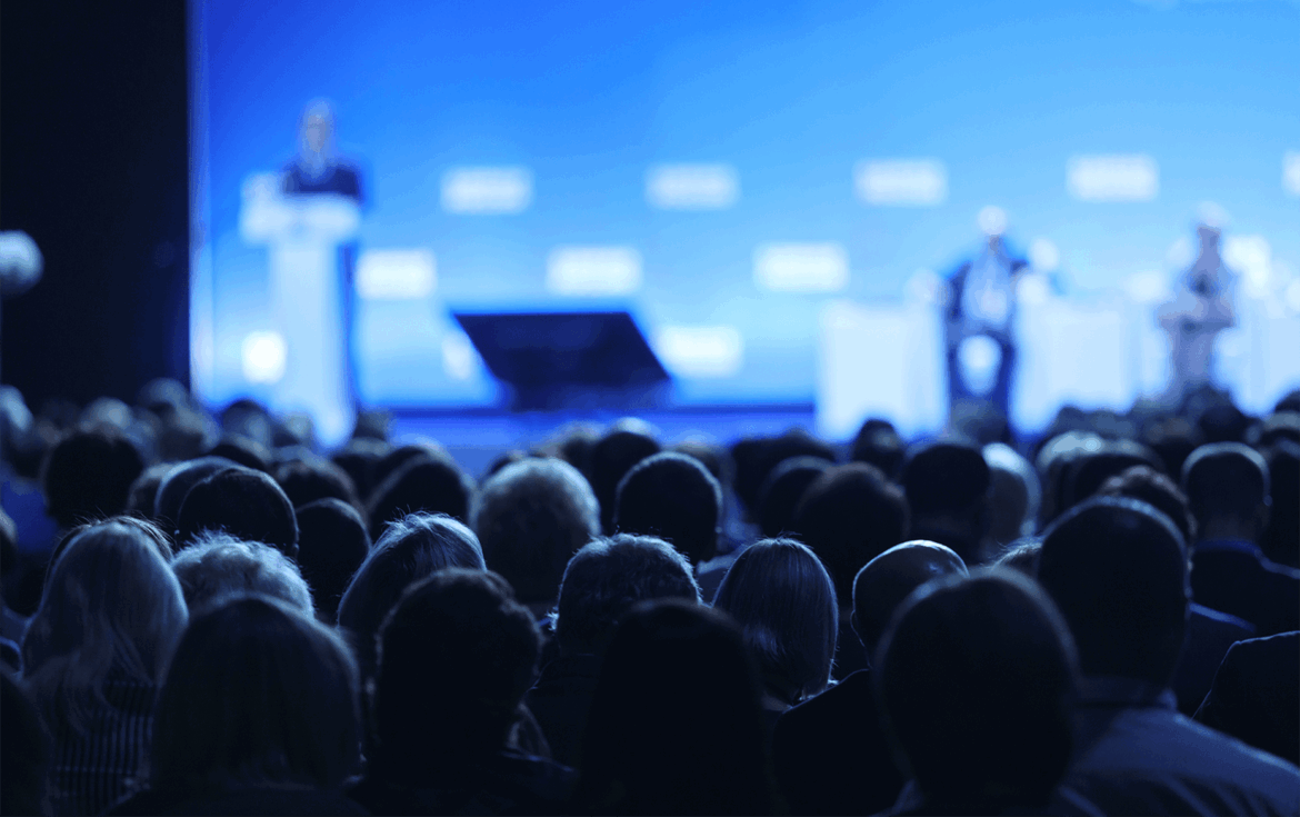 Public assistant à une conférence dans une salle plongée dans une lumière bleue, avec des intervenants sur scène en arrière-plan flou.